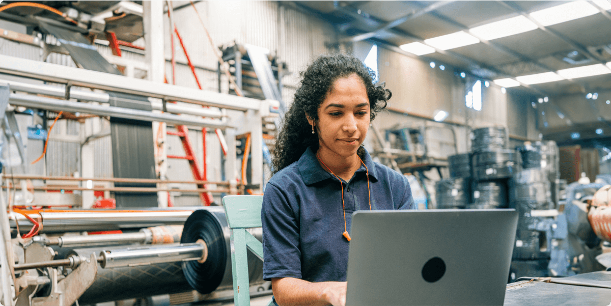 Eine Frau arbeitet an ihrem Laptop in einem Produktionsbetrieb mit Ausrüstung und Werkzeugen.